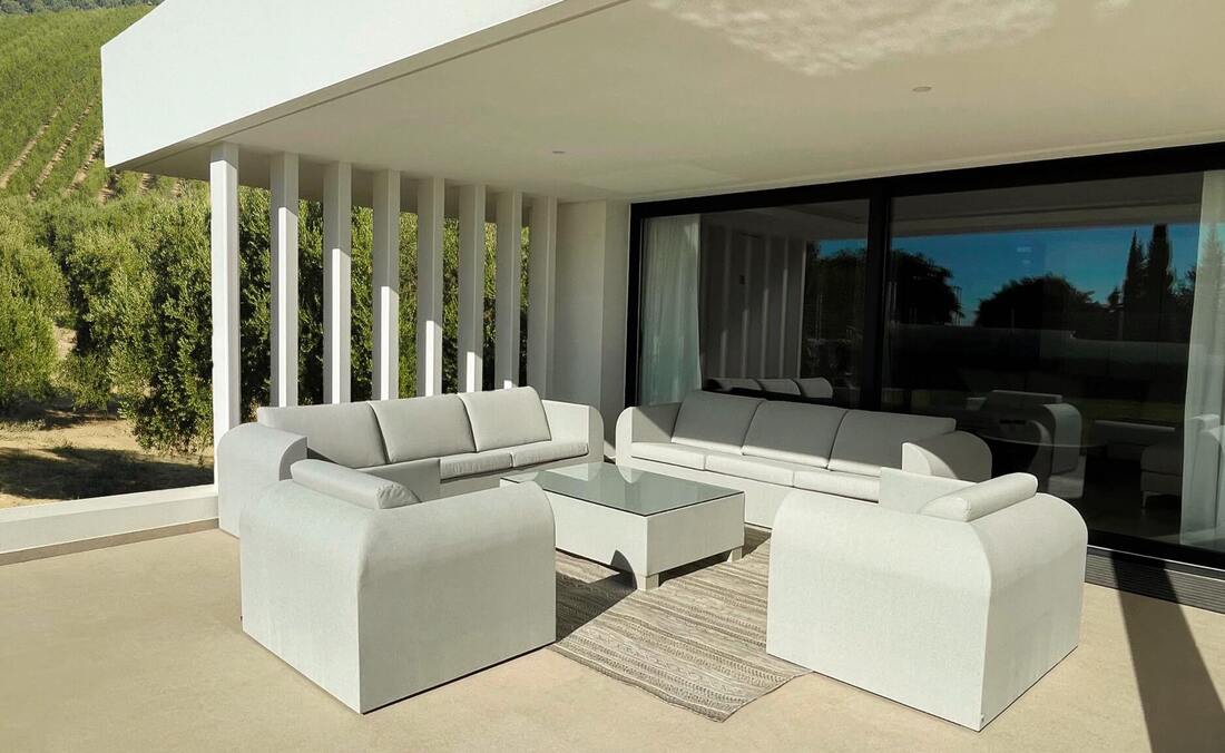 Porch in a modern Marbella house with two linen sofas, two armchairs, a coffee table and a sand-coloured outdoor carpet with olive trees field background